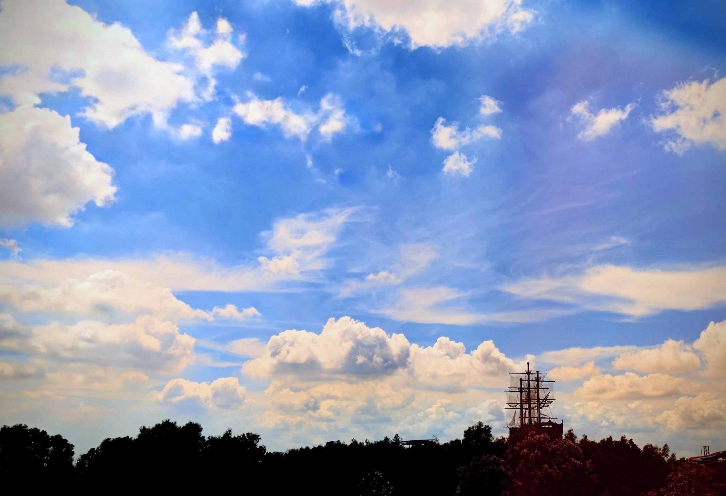 青空にわき立つ白い雲と、木々の向こうにのぞく帆船のマスト