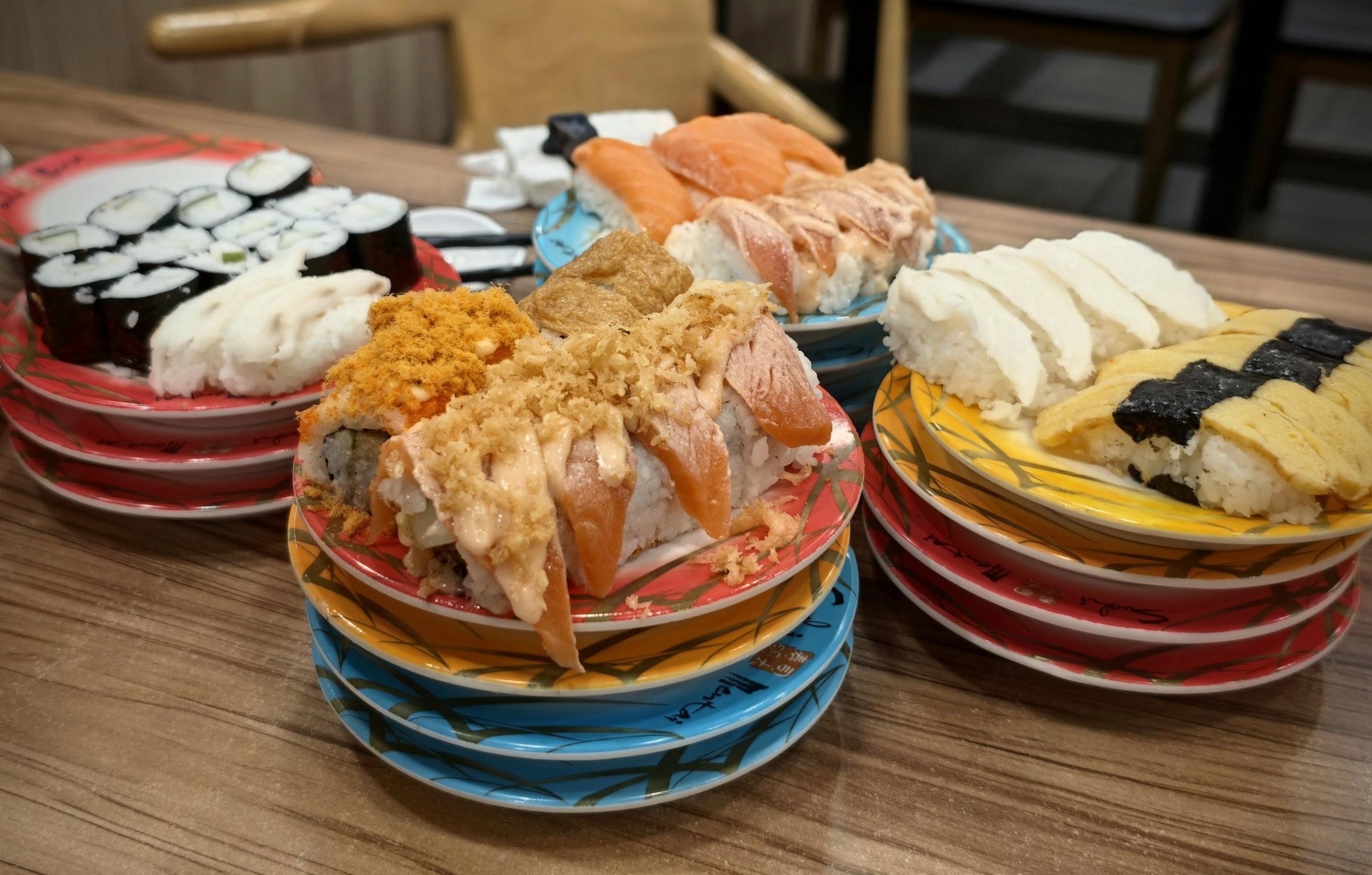 A table at a conveyor-belt sushi restaurant, plates stacked by colour: torched salmon, floss maki, tamago and kappa maki