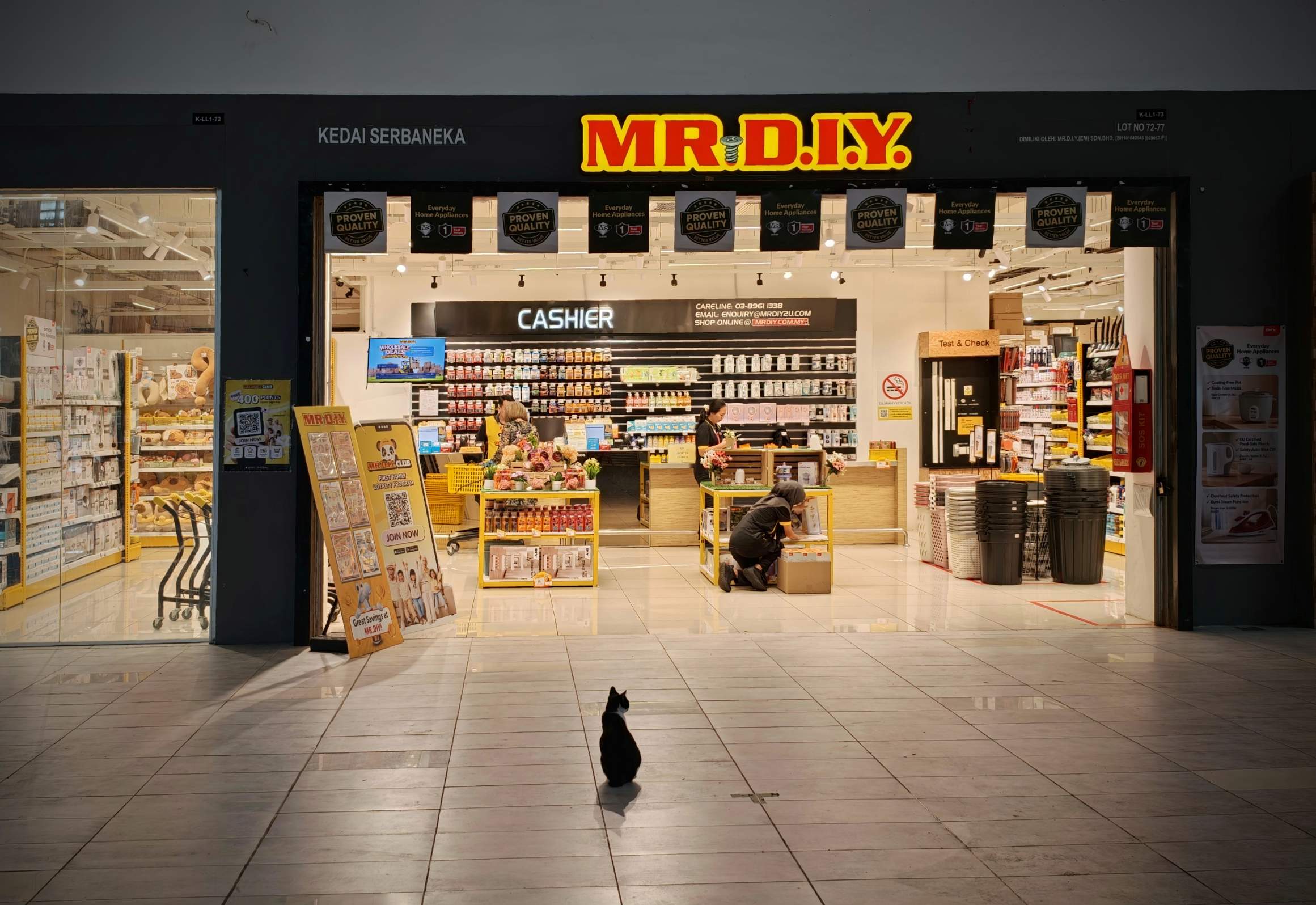 ショッピングモールの中の MR.DIY の店構え。店の前の床に黒白の猫が座っている